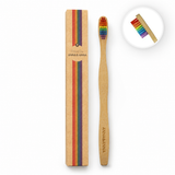 Minimalistische studiofoto van een bamboe tandenborstel met regenboogkleurige borstelharen naast een kraftkartonnen verpakking met kleurrijke strepen. Het product is gefotografeerd op een witte achtergrond met zachte professionele belichting en subtiele fotorealistische schaduwen voor een frisse, premium uitstraling.