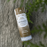 Natuurlijke sfeerfoto van een Ben & Anna Indian Mandarin deodorantstick op een verweerde houten ondergrond, omringd door zachte groene bladeren op de voorgrond. De kartonnen verpakking met botanisch dessin en aardetinten is scherp in beeld gebracht, terwijl natuurlijk licht en subtiele diepte-onscherpte zorgen voor een rustige, premium uitstraling