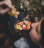 Ingezoomd sfeerbeeld van een vrouw zittend in een bosrijke omgeving, waarvan haar hoofd out of focus en afgesneden is, een lunchbox gevuld met fruit, noten en groenten heeft rusten terwijl ze een kleine tros druiven er uit haalt