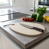 Lichte houtensnijplank van 28 x 19 cm op een licht keukenaanrecht in moderne keuken, met daarop een roestvrijstalen keukenmes. Op de achtergrond liggen verse tomaten, citroen en paprika, subtiel onscherp weergegeven in natuurlijk daglicht